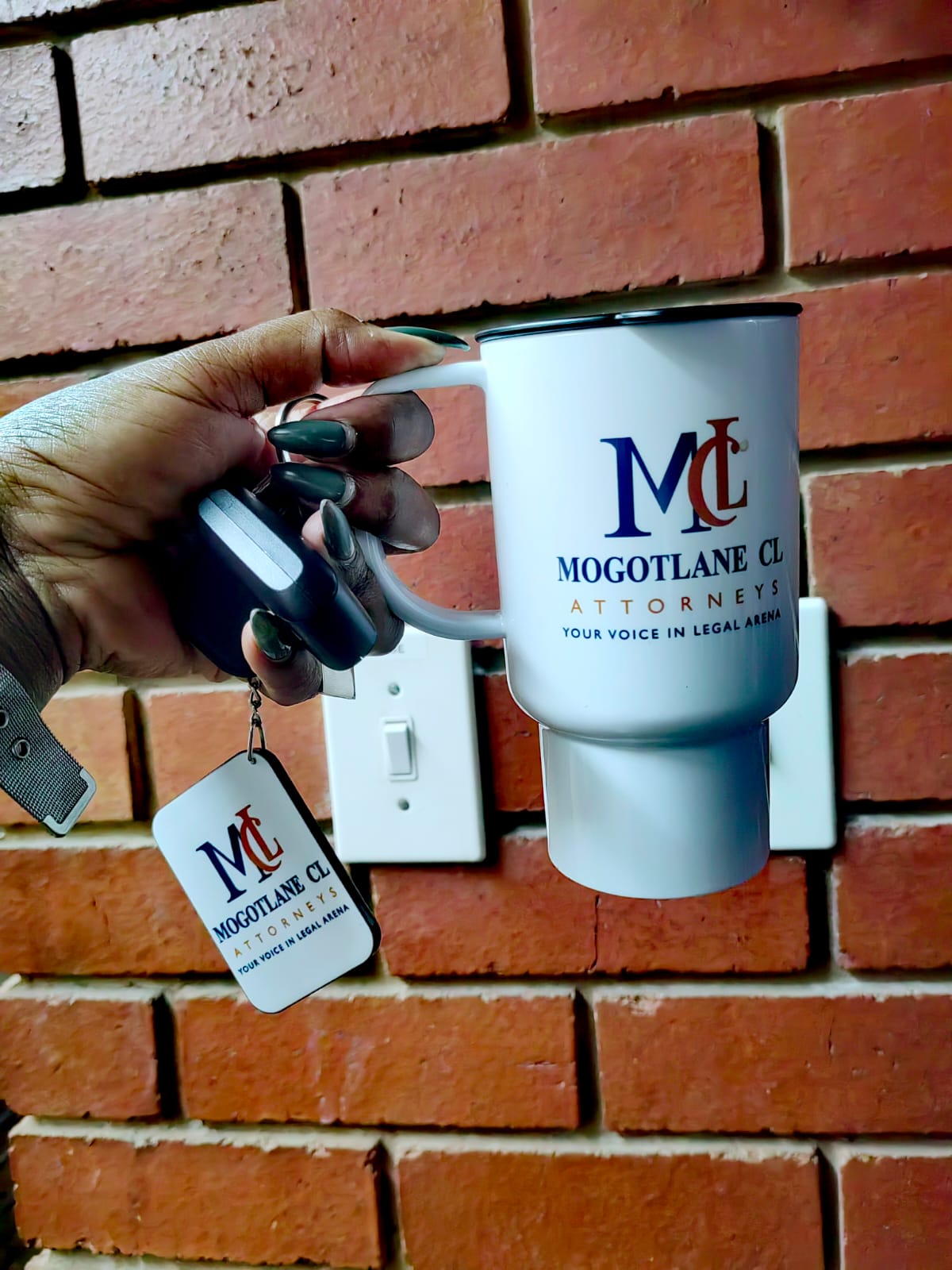 A hand holding an MCL branded travel mug next to car keys and a branded keyring